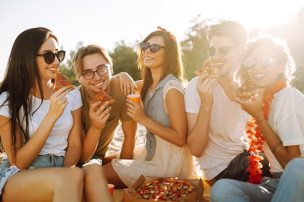 Group of young friends having picnic eating pizza toasting with beerus at the beach