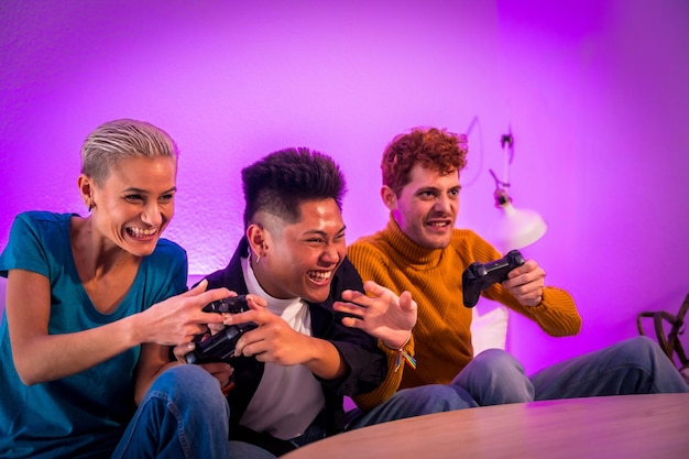 Group of young friends playing video games together on the sofa at home purple led having fun pushing