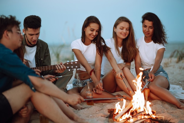 Group of young friends sitting on beach and fry sausages One man is playing guitar Camping time