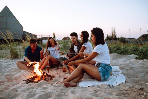 Group of young friends sitting on beach and fry sausages One man is playing guitar Camping time