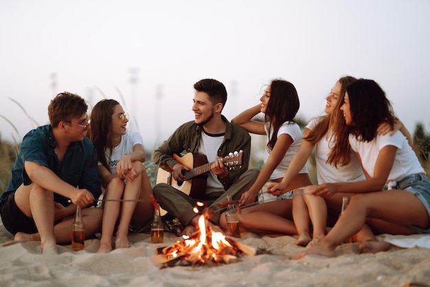 Group of young friends sitting on beach and fry sausages One man is playing guitar Camping time