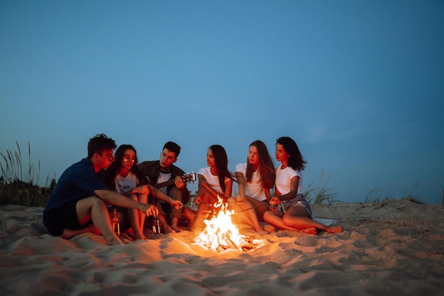 group of young friends sitting on beach and fry sausages one man is playing guitar camping time