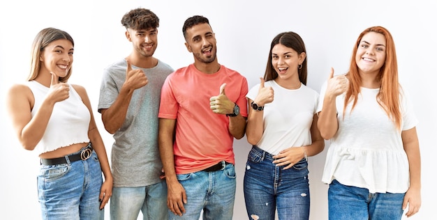 Group of young friends standing together over isolated background doing happy thumbs up gesture with hand approving expression looking at the camera showing success