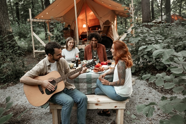 Group of young friends traveling in glamping in the forest having fun playing guitar and mbira kalimba roasting sausages sitting at dinner table near tent during summer vacation laughing hanging out