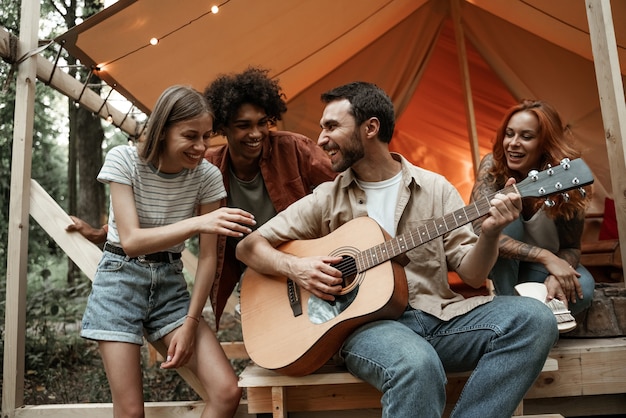 Foto gruppo di giovani amici che viaggiano in glamping nella foresta divertendosi a suonare la chitarra, mbira o kalimba e cantando canzoni arrostendo salsicce seduti in tenda durante le vacanze estive ridendo in giro