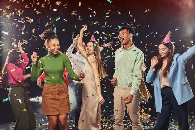 Group of young happy multiracial people wearing birthday hats dancing and having fun confetti