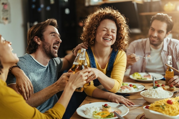 ダイニングテーブルで昼食時にビールで乾杯しながら笑って楽しんでいる若い幸せな人々のグループ