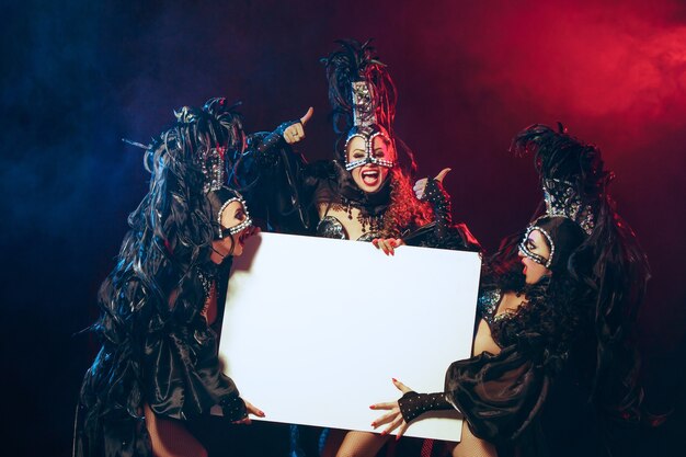 The group of young happy smiling beautiful female dancers with carnival dresses posing with empty poster on black studio background