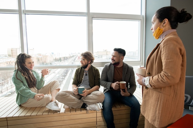 Group of young intercultural designers or managers gathered by window having coffee and discussing working plans or creative ideas at break