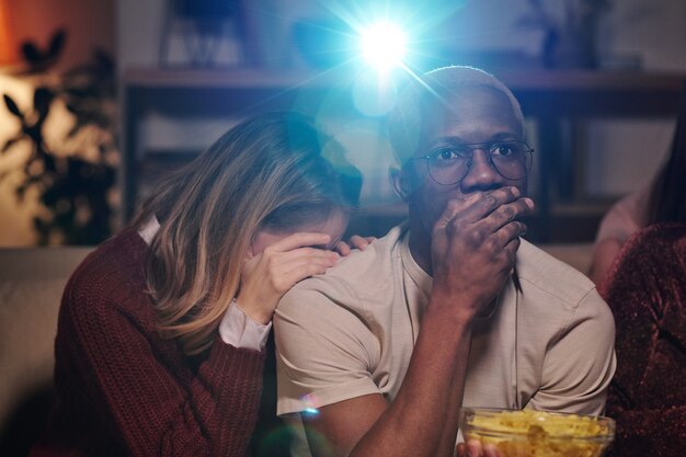 Group of young intercultural friends relaxing on couch in living-room and watching interesting movie together while enjoying home party