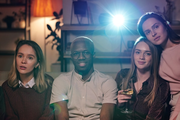 Gruppo di giovani amici interculturali che si rilassano nel soggiorno davanti alla telecamera e guardano film insieme mentre si godono la festa a casa