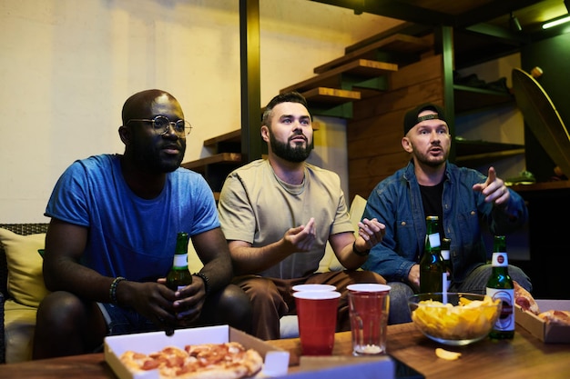 Group of young interracial buddies watching football match broadcast in garage