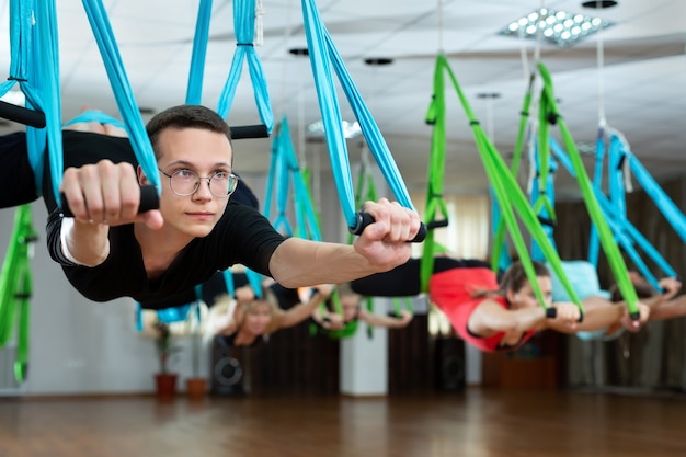 Group of young men and women do aerial yoga in hammocks at a fitness club.