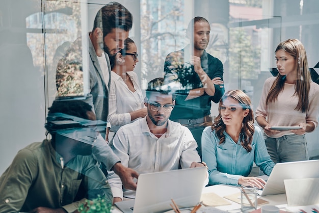 Group of young modern people in smart casual wear communicating and using modern technologies while working in the office