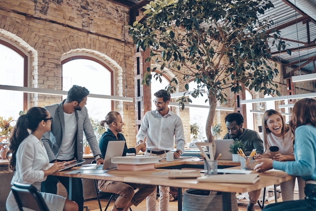 Group of young modern people in smart casual wear communicating and using modern technologies while working in the office