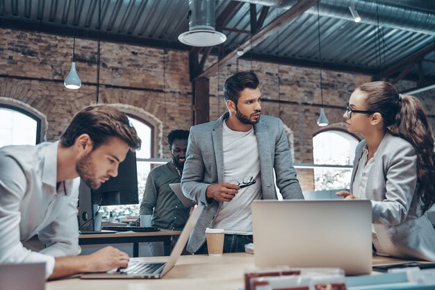 Group of young modern people in smart casual wear communicating and using modern technologies while working in the office