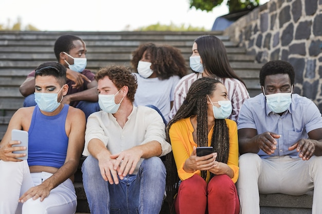 Gruppo di giovani amici multirazziali seduti sulle scale della città mentre indossano una maschera chirurgica per l'epidemia di coronavirus