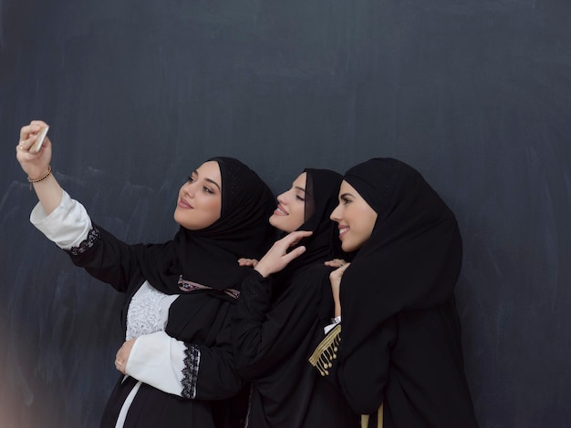 黒い背景の前で自撮り写真を撮りながら、スマートフォンを使ってヒジャーブを着たファッショナブルなドレスを着た若いイスラム教徒の女性のグループ。高品質の写真