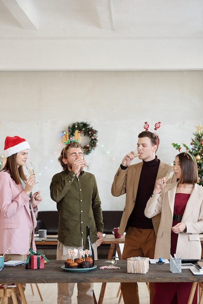 お祝いのテーブルのそばに立っている間フルートからシャンパンを飲むスマートカジュアルウェアとクリスマスのヘッドバンドの若いオフィスマネージャーのグループ