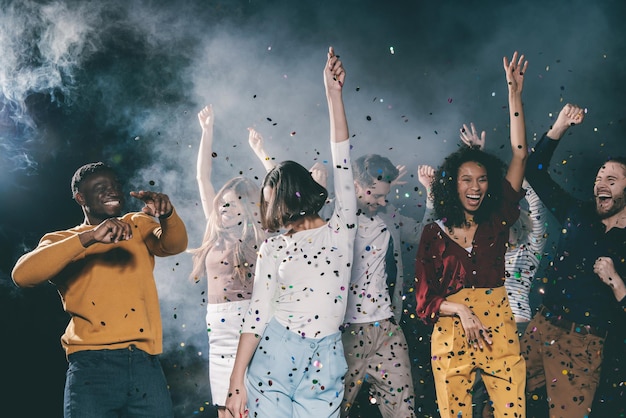Group of young people dancing and having fun in night club together while confetti flying around