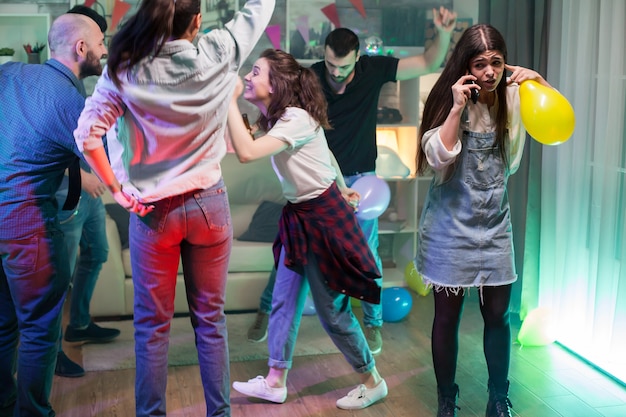 Photo group of young people dancing at a party while girl trying to have a conversation on her phone.