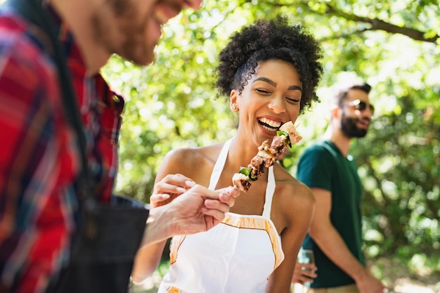 Group of young people having fun in the park at barbecue grilling and eating meat skewers together