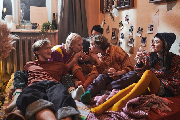 Group of young people sitting on the floor in the room smoking cigarettes and drinking alcohol drinks during domestic party