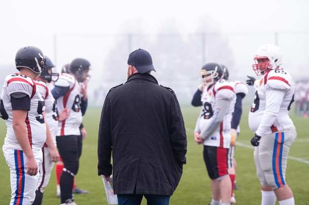 Photo group of young professional american football players with coach discussing strategy together during training match on the stadium field