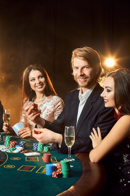 Group of young rich people is playing poker in the casino. Man in business suit and two young women in black dresses. Smoke. Casino. Poker