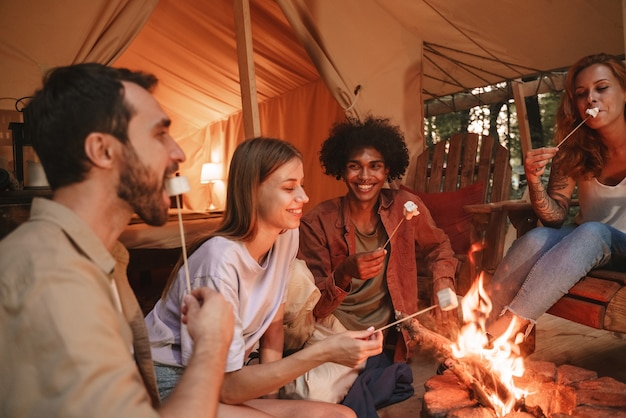 Un gruppo di giovani sorridenti che arrostiscono marshmallow su spiedini sul pozzo del fuoco al campeggio, godendosi una vacanza glamping all'aperto con gli amici insieme riaprono dopo il blocco della pandemia.