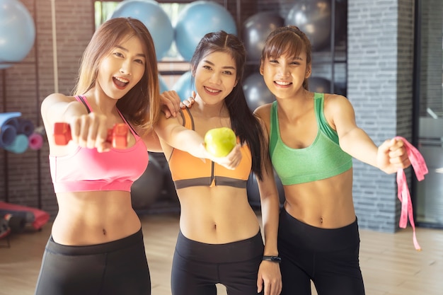 Group of young sport women in gym, showing dumbbell, green apple and tape