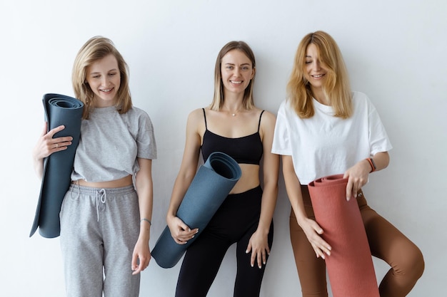 A group of young sports girls with yoga mats standing against a white wall Girlfriends in the gym relaxing after fitness or yoga indoors