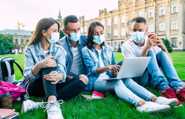 Un gruppo di giovani studenti in maschere mediche con laptop e libri stanno studiando insieme all'università. amici all'aperto seduti sull'erba.