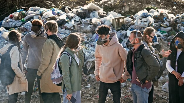 Group of young volunteers in gas masks take care of the\
environment while standing at garbage dumpac...