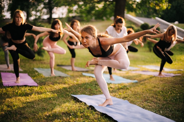 スポーツウェアの若い女性のグループは、日の出の間に公園で片足の朝にヨガの位置の1つに立っています。瞑想する人々のグループ。太陽光線がカメラに輝いています