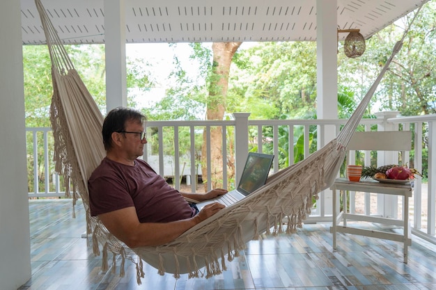 Grown man rests and working a laptop in a hammock on a terrace home near tropical garden Thailand