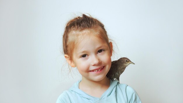 Grown up starling nestling sit on shoulder of little girl and sing with open beak Portrait of happy child on gray