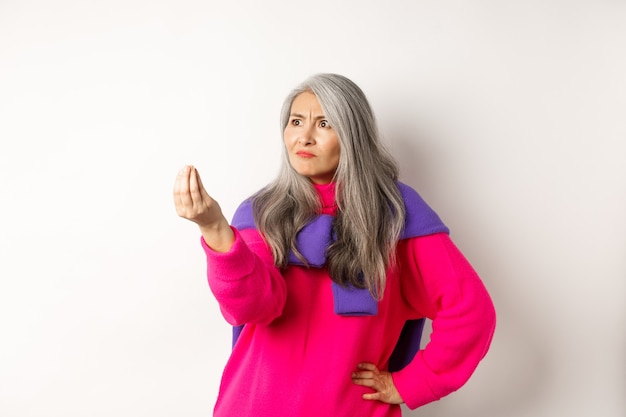 Grumpy asian grandmother complaining, showing italian what do you want gesture, shaking fingers and staring angry left at copy space, standing over white background