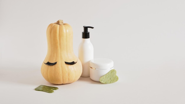 Gua sha natural jadeite stone scrapers, cream jar and bottle,
pumpkin with false eyelashes gray background, face peeling with
massage