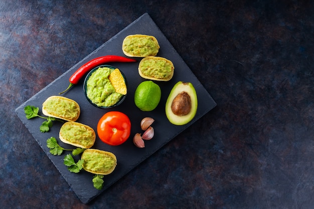 Guacamole sauce, nachos chips and ingredients on a dark background. Mexican guacamole sauce with avocado, ingredients and corn nachos. Copy space. Top view