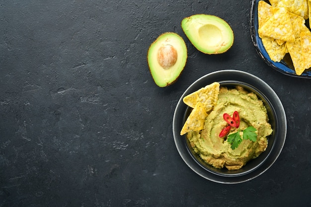 Guacamole. Traditional latinamerican Mexican dip sauce in a black bowl with avocado and ingredients and corn nachos. Avocado spread. Top view. Copyspace.