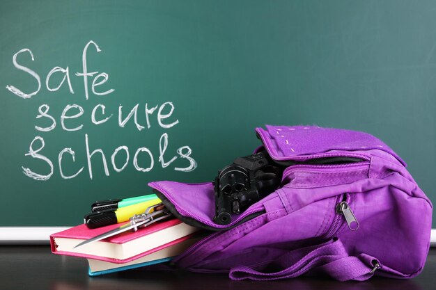 Gun in school backpack on wooden desk on blackboard background