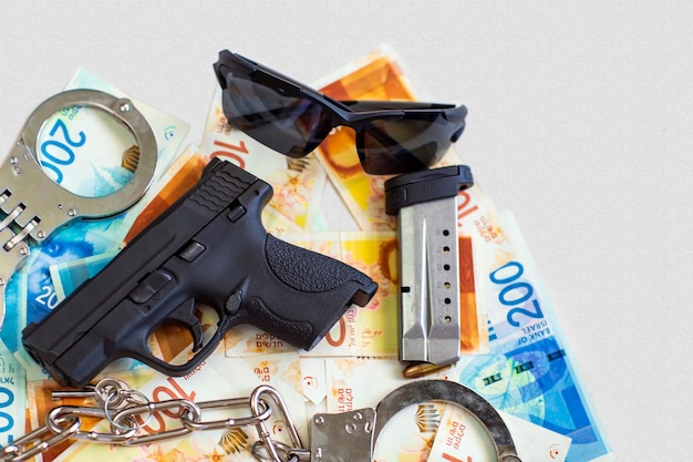 Pistola, due manette della polizia, occhiali da sole sullo sfondo delle banconote del nuovo shekel israeliano. pistola semiautomatica arma da fuoco con caricatore su shekel con le nuove banconote da 100, 200 nis, valuta. corruzione