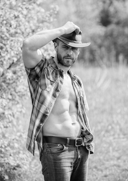 Guy bearded cowboy in nature Macho six packs torso wear rustic style clothes and cowboy hat Strong and confident cowboy Owner of rancho Man unshaven face muscular torso cowboy Farm concept