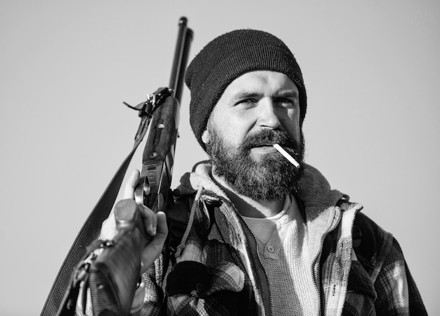 Guy bearded hunter spend leisure hunting and smoking hunting\
masculine hobby concept man brutal bearded guy gamekeeper blue sky\
background brutality and masculinity hunter with rifle gun close\
up