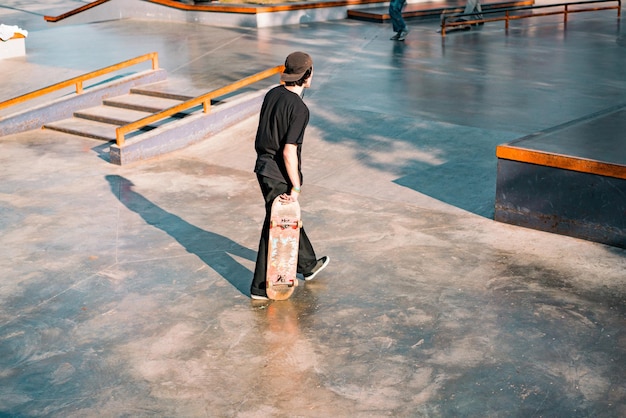 A guy in a black clothes and sneakers walks on a skate park.\
sport. style. leisure. city. urban. outdoor. scratched used\
skateboard. different shapes. stairs. blocks. concrete. lifestyle.\
skate-park