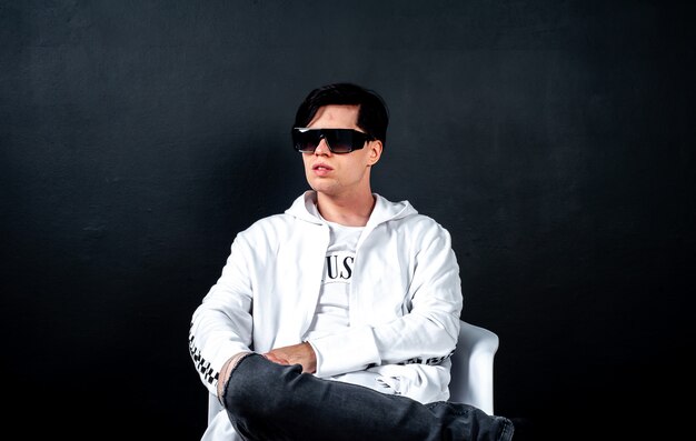 a guy in black glasses is sitting on a chair against a dark background of concrete in a studio; in the studio is a young guy in black glasses