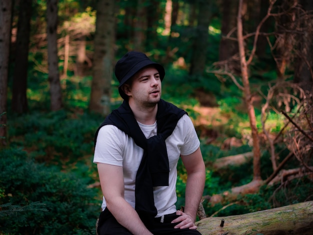 A guy in a black hat and a white T-shirt sits on a log in the forest and smiles