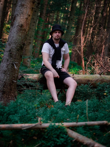 A guy in a black hat and a white T-shirt sits on a log in the forest and smiles