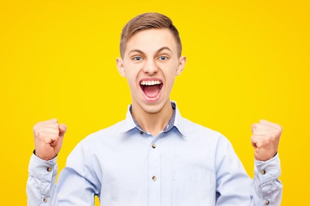 Guy in blue shirt rejoices victory isolated on yellow background, raised hands up